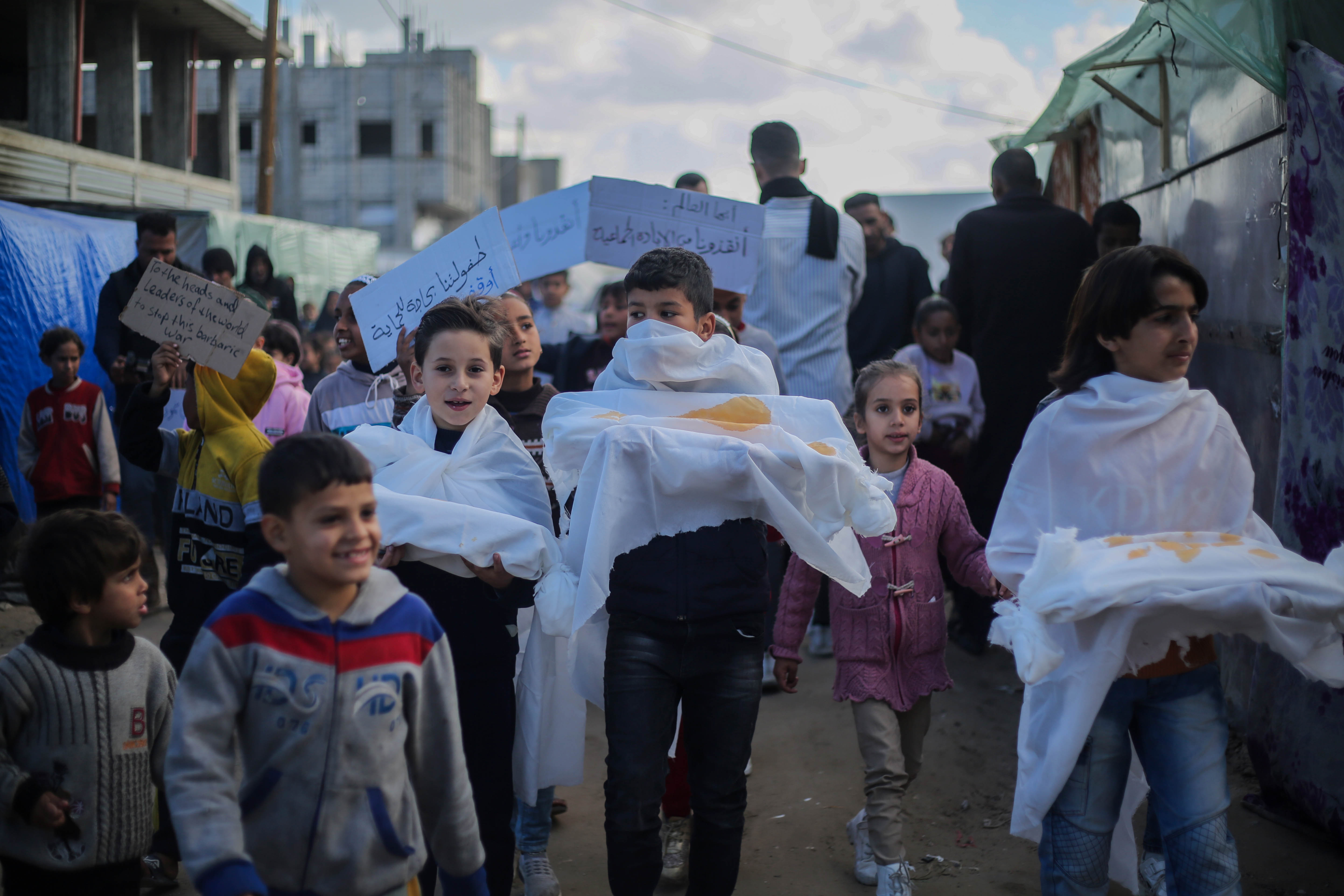 children hold pretend bodies wrapped in white sheets during a protest at a refugee camp